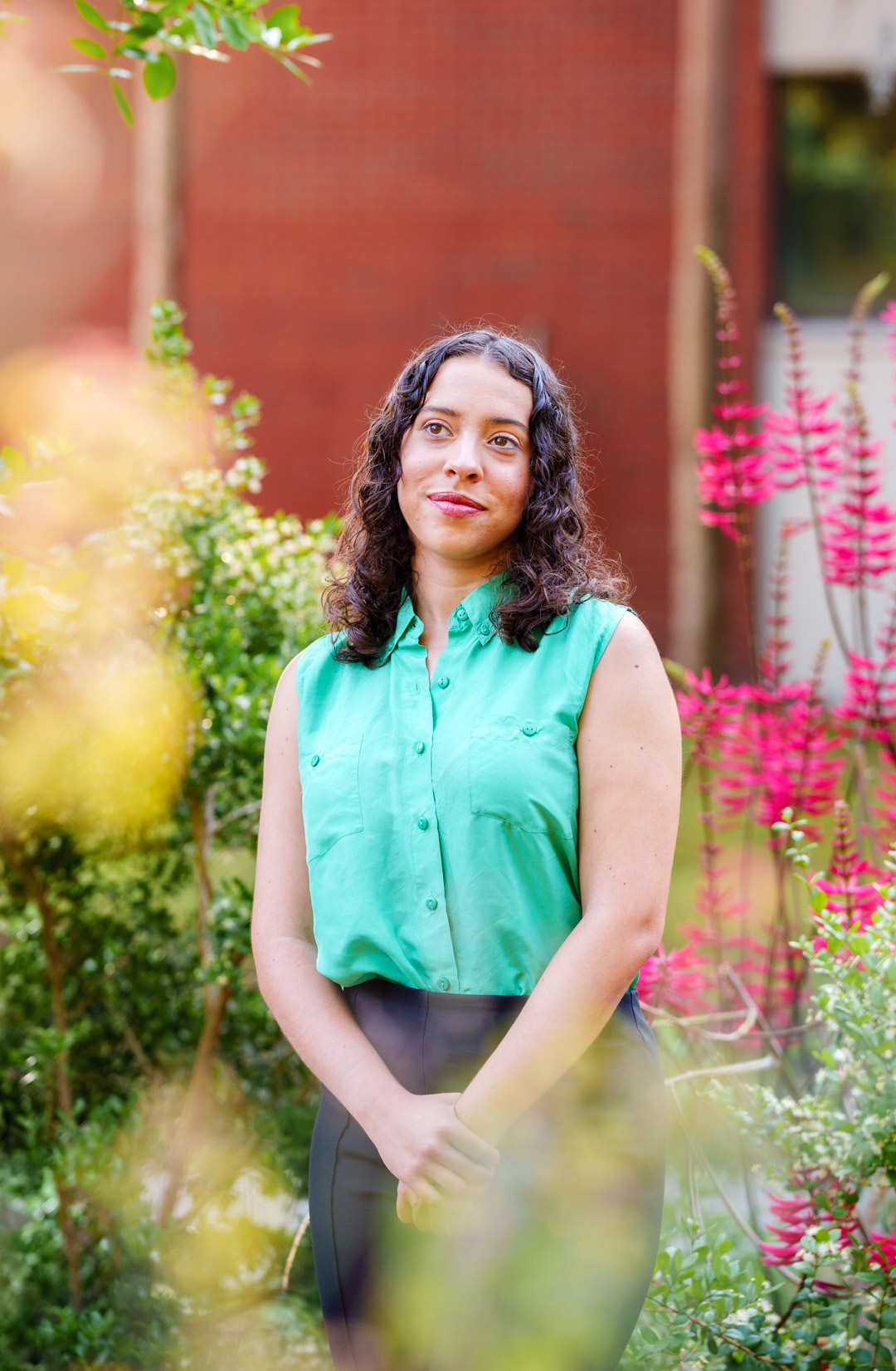 An outdoors portrait of Natalie Triana in flowers, with a red brick wall in the background.