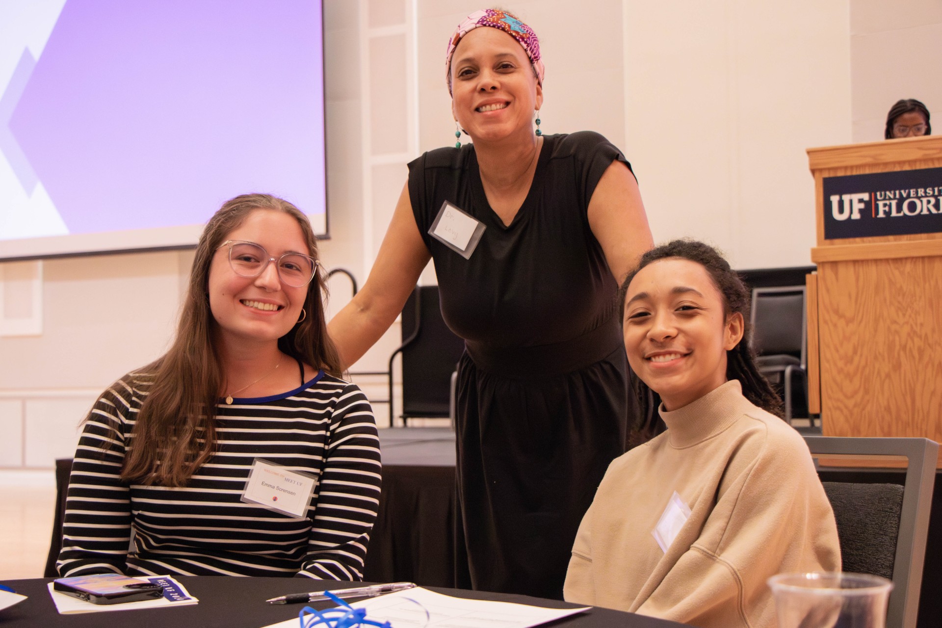 Three women, two seated and one standing behind, smile at the camera.